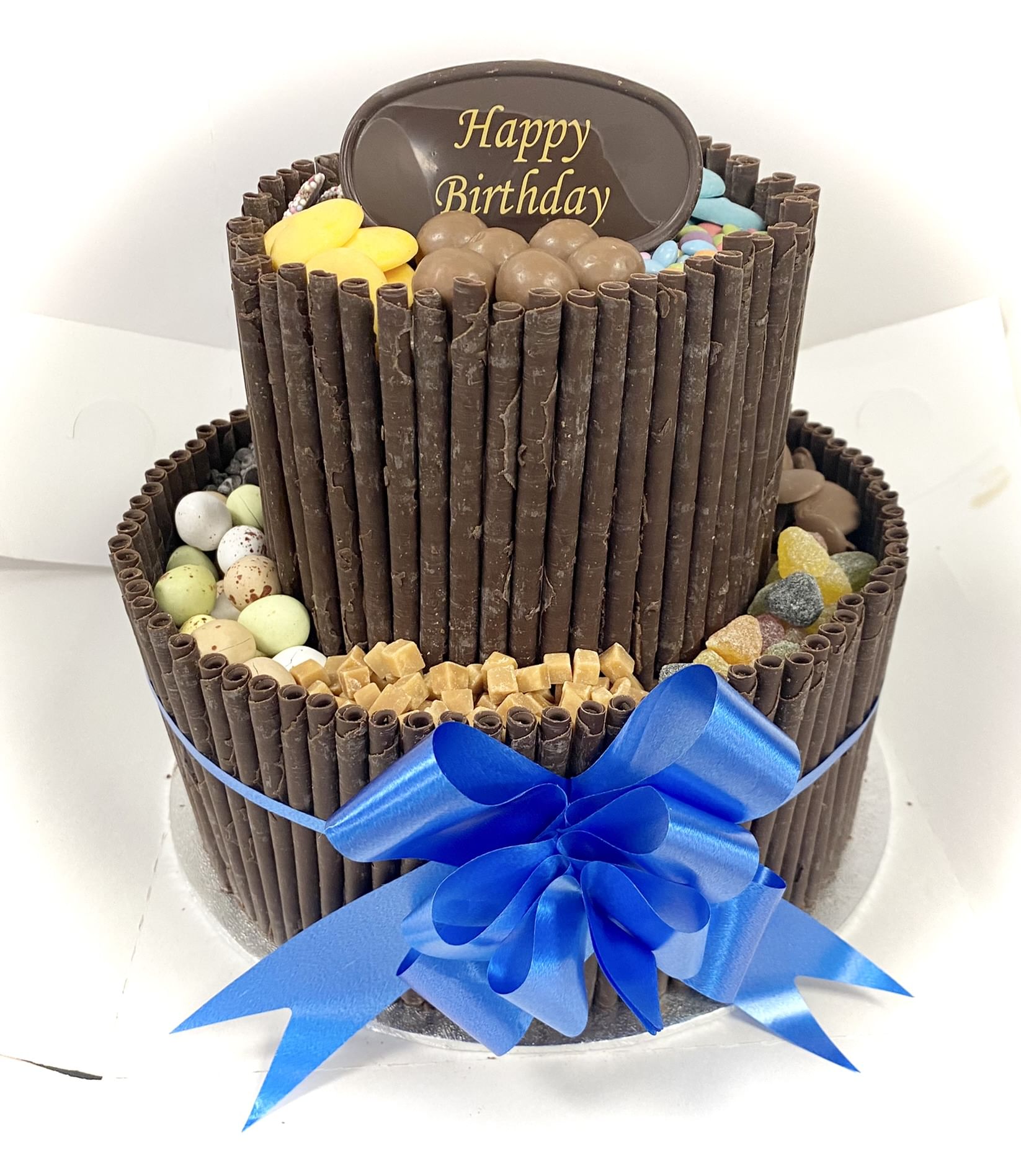 A two-tiered Chocolate Blackout Cake decorated with chocolate wafer rolls around the sides. The cake is adorned with various candies, including chocolate pieces and colorful candy eggs. A blue ribbon is tied around the bottom tier, and a 'Happy Birthday' plaque tops the cake.